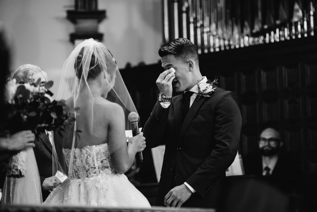 Photo of a couple, bride holds microphone, groom wipes tears from his eyes